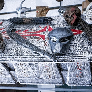 May include: A display of Predator-themed collectibles, including a skull, mask, and bladed weapon, arranged on a textured, gray surface with red accents. The items are detailed and appear to be replicas.