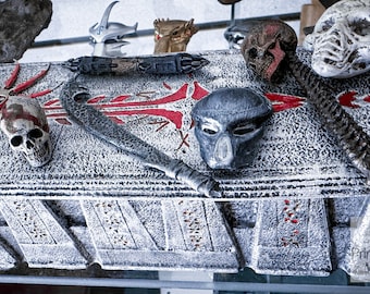 May include: A display of Predator-themed collectibles, including a skull, mask, and bladed weapon, arranged on a textured, gray surface with red accents. The items are detailed and appear to be replicas.