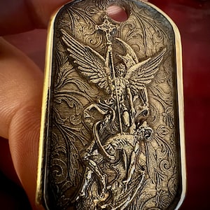 May include: A gold-coloured pendant featuring an engraved image of an angel with wings, holding a staff, and defeating a demon. The pendant has a decorative floral pattern background and a hole at the top.