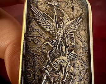 May include: A detailed, gold-toned pendant with an engraved image of an angel with wings, holding a staff, and defeating a demon. The pendant has a decorative floral pattern background and a hole at the top.