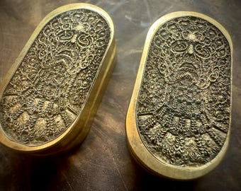 May include: Two brass-colored, rectangular boxes with rounded edges. Each box features a detailed, embossed design of a face with intricate patterns. The boxes are displayed on a dark, textured surface, showcasing the metallic sheen.