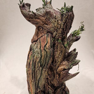 May include: A detailed sculpture of a tree-like figure with a textured, weathered appearance. The sculpture features shades of brown, green, and gray, with intricate details mimicking bark, moss, and foliage. The base is a gray stone-like structure.