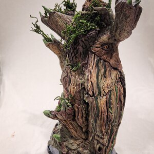May include: A detailed, hand-painted sculpture of a tree with a face, featuring textured bark in shades of brown, green, and gray. The tree has multiple branches with green leaves and moss. The base is a gray stone.