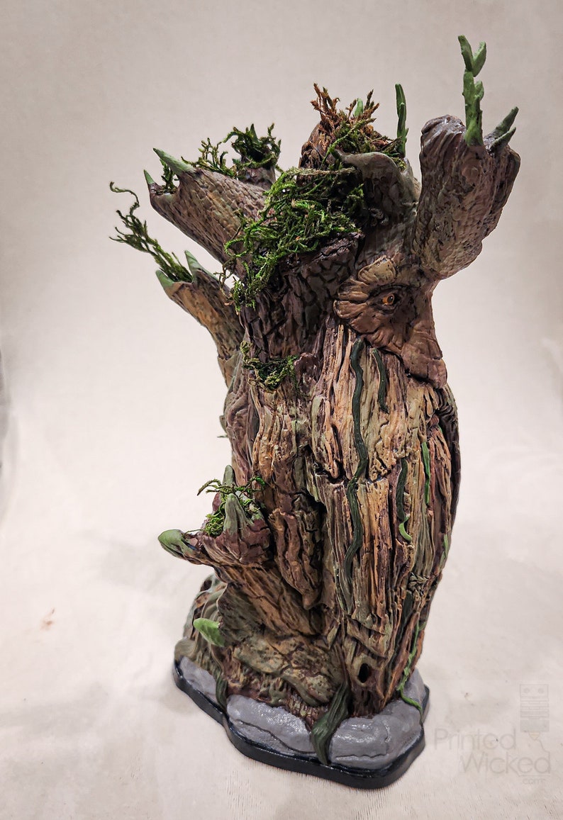 May include: A detailed, hand-painted sculpture of a tree with a face, featuring textured bark in shades of brown, green, and gray. The tree has multiple branches with green leaves and moss. The base is a gray stone.