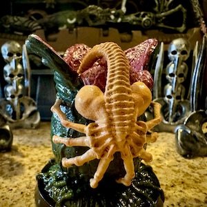 May include: A detailed sculpture of a facehugger, a creature from the Alien film franchise, in a menacing pose. The facehugger is tan and brown, with a long tail and spider-like legs. The sculpture is set against a backdrop of skull-shaped decorations.