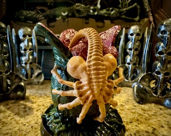 May include: A detailed sculpture of a facehugger, a creature from the Alien film franchise, in a menacing pose. The facehugger is tan and brown, with a long tail and spider-like legs. The sculpture is set against a backdrop of skull-shaped decorations.
