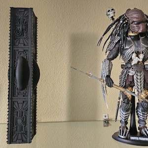 May include: A black, rectangular, intricately designed sculpture stands next to a detailed action figure of a Predator. The Predator figure is in a battle-ready pose, with a spear and other weapons. The figure is on a black stand.