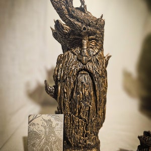 May include: A bronze-colored sculpture of a Green Man figure with a long beard and leafy crown. The sculpture has a textured surface and is next to a small card with a similar illustration and the year 2023.