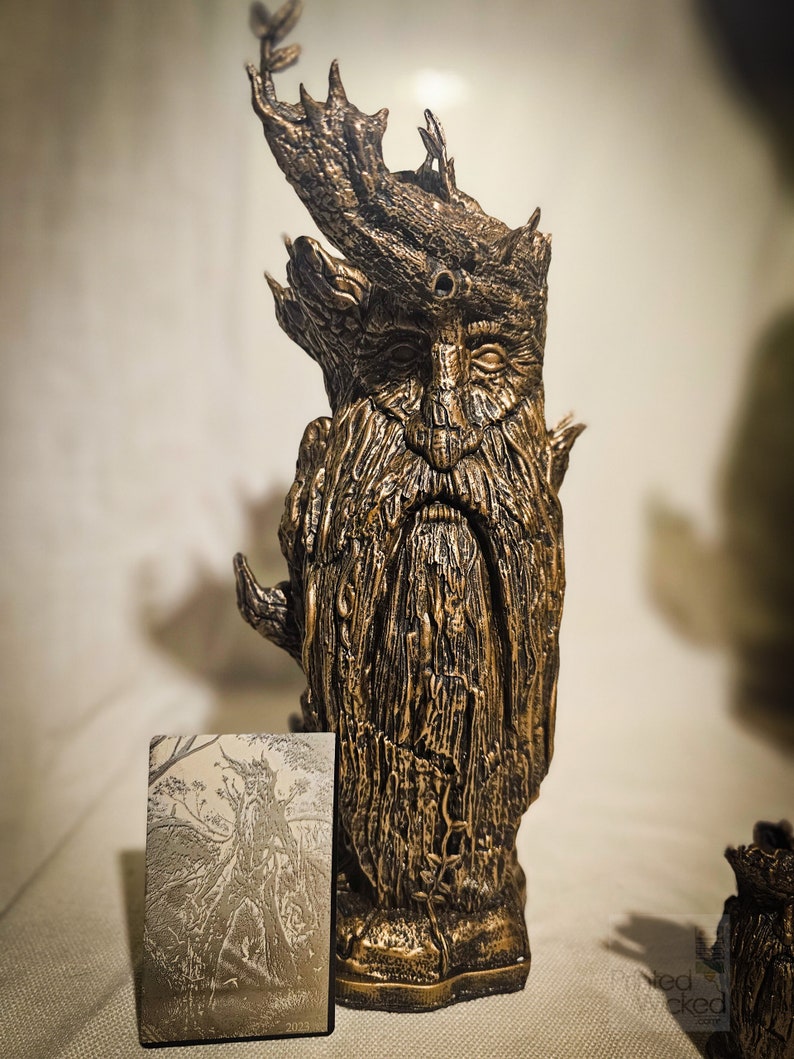 May include: A bronze-colored sculpture of a Green Man figure with a long beard and leafy crown. The sculpture has a textured surface and is next to a small card with a similar illustration and the year 2023.