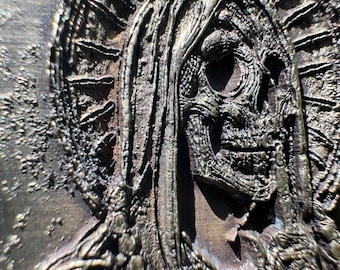 May include: Close-up of a bronze-colored relief sculpture depicting a skeletal figure with a halo. The figure's face is detailed with hollow eye sockets and a visible skull. The sculpture has a textured surface and appears aged.