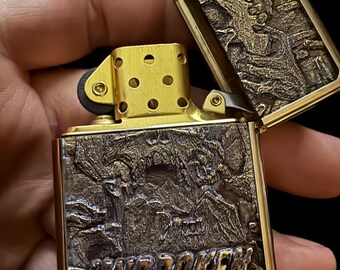 May include: A brass-colored lighter with an open lid, revealing an engraved design. The lighter's body features a detailed landscape scene with the word "UNBROKEN" at the bottom. The lid also has an engraved design. The lighter is held in a hand.