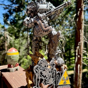 May include: A detailed figurine of a silver-colored armored figure holding a large gun. The figure stands on a rocky base with a yellow and black danger sign. A miniature bomb is visible in the background.