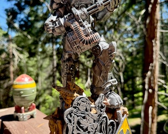 May include: A detailed figurine of a silver-colored armored figure holding a large gun. The figure stands on a rocky base with a yellow and black danger sign. A miniature bomb is visible in the background.