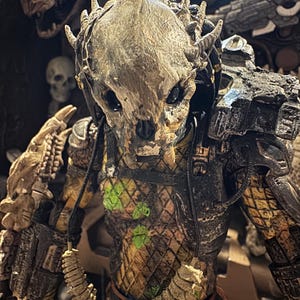 May include: A detailed action figure of a Predator alien, featuring a textured, bone-colored mask and intricate armor. The figure has a detailed chest with green accents and a brown and gold color scheme. The figure is a collectible item.