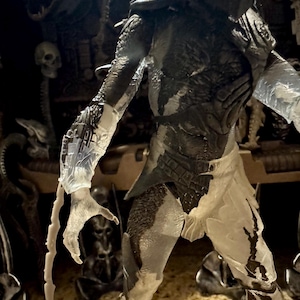 May include: A detailed action figure of a Predator, featuring a dark gray and translucent design. The figure has intricate armor, a helmet, and extended claws. The figure is posed in a dynamic stance, showcasing its detailed textures and design.