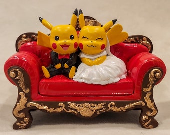 May include: A figurine of two Pikachu characters dressed as a bride and groom, seated on a red, ornate sofa. The sofa has gold accents and a dark brown frame. The bride Pikachu wears a white dress and veil, while the groom wears a black tuxedo.