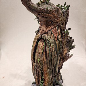 May include: A detailed sculpture of a tree-like figure with a long beard and leafy crown. The figure has a weathered, textured appearance with shades of brown, green, and gray. The base is a gray stone-like structure.
