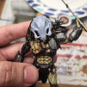 Op de afbeelding: Gedetailleerde actiefiguur van een Predator, met een lichtgrijs masker, ingewikkelde bepantsering en een speer. De figuur is beschilderd met een combinatie van zwart, zilver en bruin, wat het iconische ontwerp van het personage laat zien.