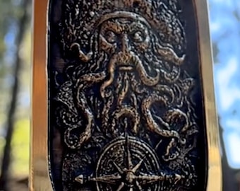 May include: Oval pendant with a detailed relief of a bearded sea god wearing a crown, with tentacles and a compass rose. The pendant has a gold-colored border and a dark, textured background. The design is intricate and maritime-themed.
