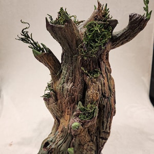 May include: A detailed, hand-painted tree sculpture with a textured, brown bark. Green moss and small plants are nestled in the crevices and branches. The base is dark with a stone-like texture, creating a fantasy or woodland theme.
