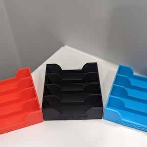Puede incluir: Tres organizadores de almacenamiento de varios niveles en rojo, negro y azul. Cada organizador tiene una base rectangular con cortes curvos y el texto "Crypto Cave" en la parte delantera. Los organizadores están diseñados para guardar y organizar artículos.