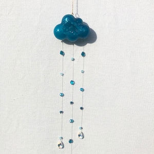 Peut inclure: Un nuage en résine bleu sarcelle avec une chaîne dorée suspendue. Le nuage a une finition scintillante et est décoré de perles de cristal bleues et transparentes et de cristaux en forme de goutte.