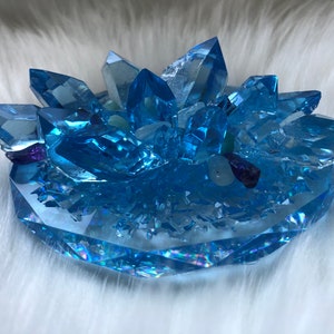 May include: A blue crystal cluster sculpture with a faceted base. The sculpture features multiple crystal points and smaller stones in shades of blue, purple, and green. The crystals are arranged on a clear base.