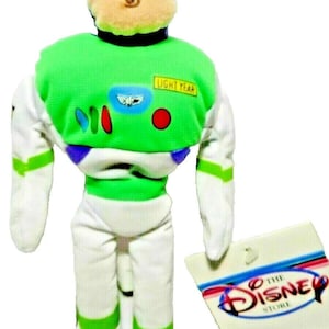 May include: A plush Buzz Lightyear doll from the Disney Store. The doll features a green and white spacesuit with a green chest and accents, and a tan head. The tag reads "The Disney Store".