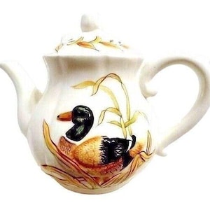 May include: Cream-coloured ceramic teapot with a duck illustration. The duck has black, brown, and orange feathers, set against yellow reeds. The lid and handle are also cream, creating a decorative and functional item.