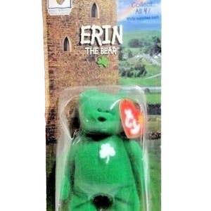 May include: A green plush bear toy named "Erin" with a white shamrock on its chest, packaged in a clear plastic case. The packaging features a castle illustration and the text "Erin The Bear". A red heart-shaped tag is attached.