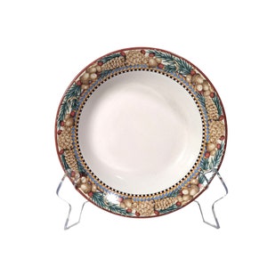 May include: A white ceramic bowl with a decorative rim featuring pinecones, acorns, and berries. The rim has a red border, a blue and gold geometric pattern, and a black and white checkered band. The bowl is displayed on a clear plastic stand.