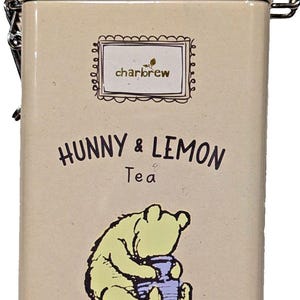 Puede incluir: Una lata de té rectangular con fondo beige claro y tapa azul claro. La lata presenta el texto "Hunny & Lemon Tea" y una ilustración de un oso amarillo. Contiene 60 bolsitas de té y pesa 120g.