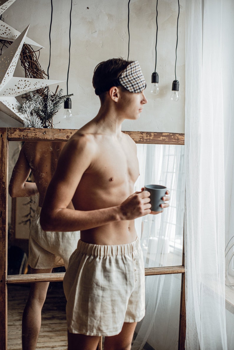 Op de afbeelding: Een persoon met een slaapmasker en een witte boxershort staat voor een spiegel en houdt een grijze mok vast. De persoon kijkt uit het raam.
