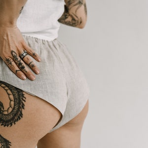Pu&ograve; includere: Pantaloncini in lino beige chiaro con elastico in vita. Gli shorts sono abbinati a un top bianco. L'immagine mostra una persona con tatuaggi sul braccio, sulla mano e sulla gamba. I tatuaggi sono neri e dettagliati.