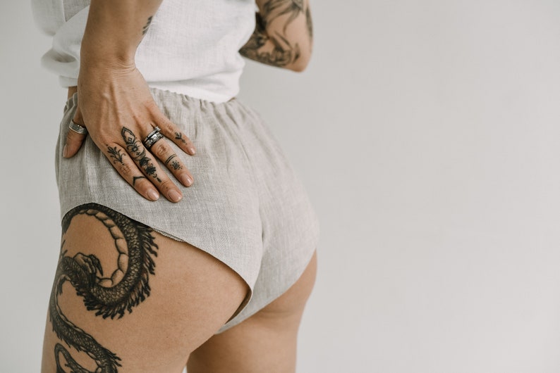 Pu&ograve; includere: Pantaloncini in lino beige chiaro con elastico in vita. Gli shorts sono abbinati a un top bianco. L'immagine mostra una persona con tatuaggi sul braccio, sulla mano e sulla gamba. I tatuaggi sono neri e dettagliati.