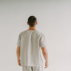 May include: A light beige linen shirt with short sleeves. The shirt has a relaxed fit and a minimalist design. The model is wearing matching shorts. The outfit is suitable for casual wear.