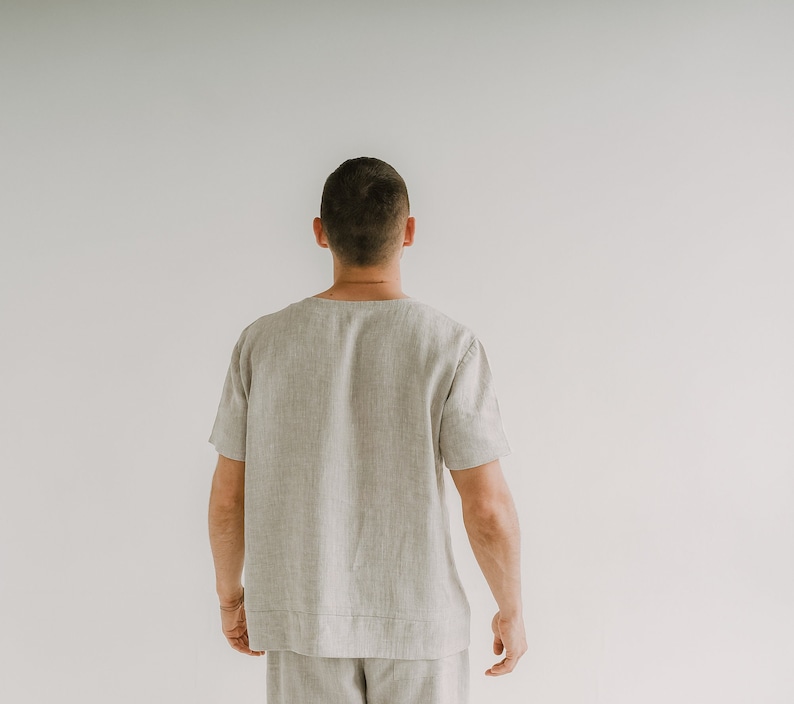 May include: A light beige linen shirt with short sleeves. The shirt has a relaxed fit and a minimalist design. The model is wearing matching shorts. The outfit is suitable for casual wear.