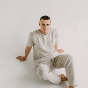 May include: A light grey linen short-sleeved shirt and matching trousers set. The casual outfit features a crew neck shirt and drawstring trousers. The model is sitting on a white surface.