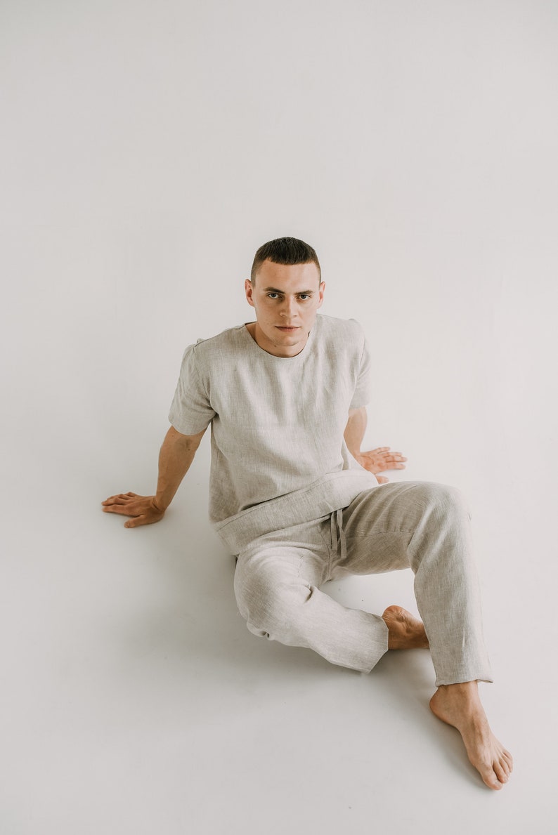 May include: A light grey linen short-sleeved shirt and matching trousers set. The casual outfit features a crew neck shirt and drawstring trousers. The model is sitting on a white surface.