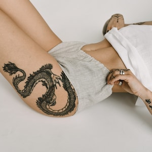 Pu&ograve; includere: Una persona indossa pantaloncini grigio chiaro e un top bianco. Un grande tatuaggio di drago nero &egrave; visibile sulla coscia. La persona &egrave; sdraiata su una superficie bianca.