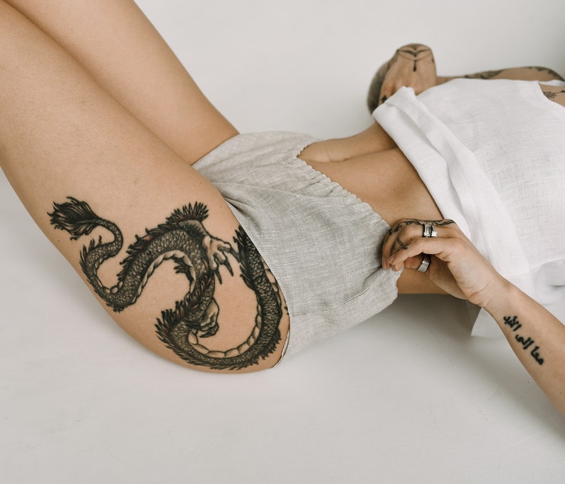 Pu&ograve; includere: Una persona indossa pantaloncini grigio chiaro e un top bianco. Un grande tatuaggio di drago nero &egrave; visibile sulla coscia. La persona &egrave; sdraiata su una superficie bianca.