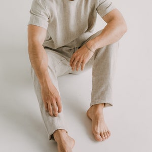 Op de afbeelding: Een persoon draagt een lichtbeige linnen shirt en een bijpassende broek. De persoon zit met gekruiste benen en blote voeten.