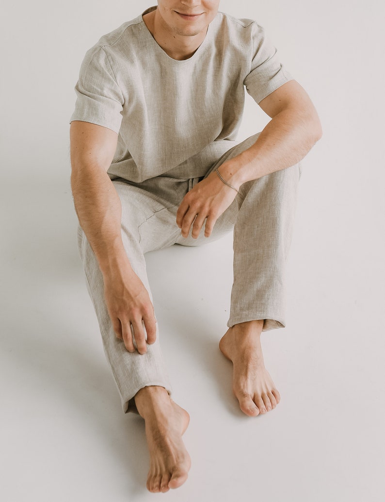 Op de afbeelding: Een persoon draagt een lichtbeige linnen shirt en een bijpassende broek. De persoon zit met gekruiste benen en blote voeten.