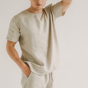 May include: A light beige linen t-shirt with short sleeves and a crew neck. The matching linen trousers have a drawstring waist. The outfit is made of a lightweight, breathable fabric, ideal for warm weather. The clothing is a neutral colour.
