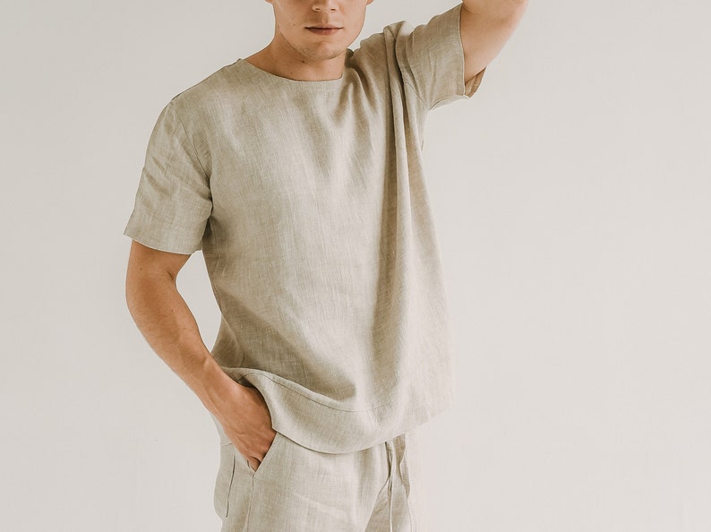 May include: A light beige linen t-shirt with short sleeves and a crew neck. The matching linen trousers have a drawstring waist. The outfit is made of a lightweight, breathable fabric, ideal for warm weather. The clothing is a neutral colour.