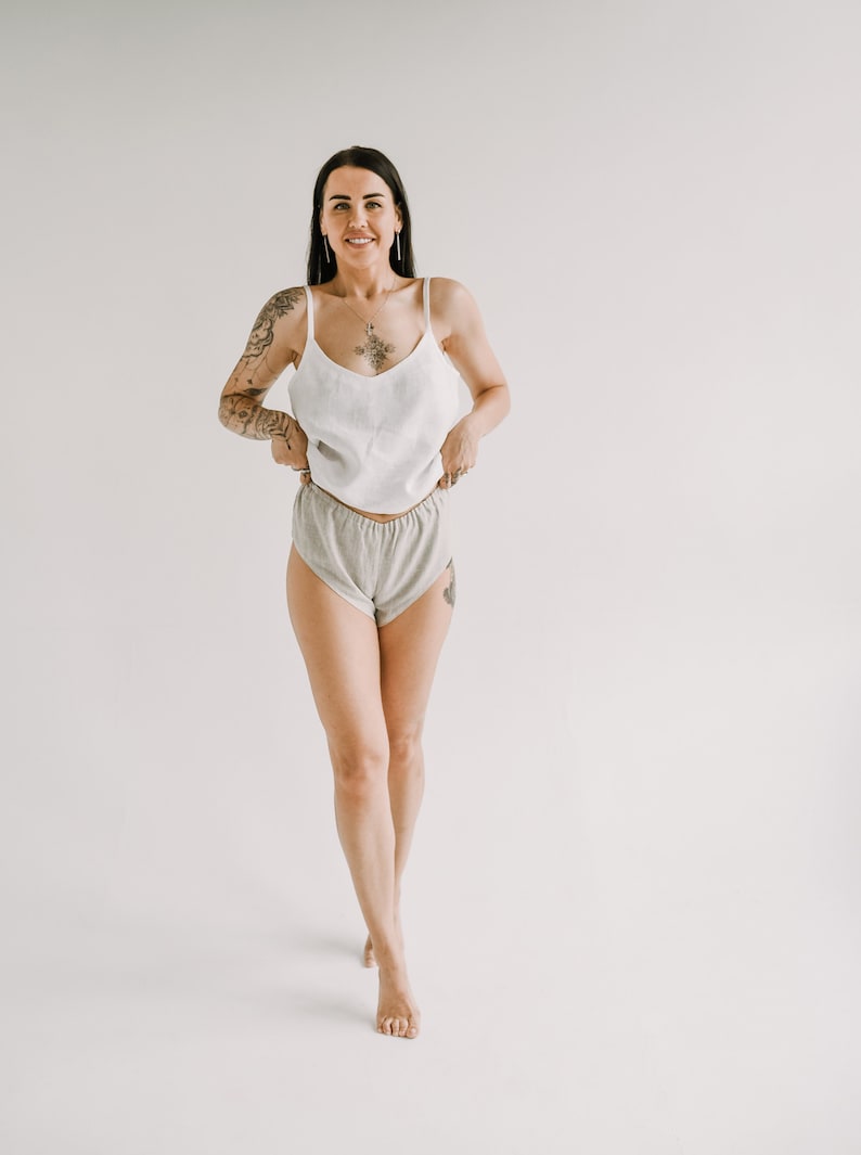 Pu&ograve; includere: Una donna indossa un top bianco con spalline e pantaloncini grigi. Il top ha spalline sottili e scollo a V. I pantaloncini sono a vita alta e realizzati in un tessuto morbido e chiaro. La donna sorride davanti a uno sfondo bianco.
