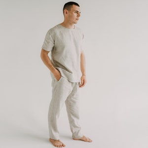 Op de afbeelding: Een persoon draagt een lichtbeige linnen shirt en een bijpassende broek. Het shirt heeft korte mouwen en een ronde hals. De broek heeft een trekkoord in de taille en is losjes gesneden.