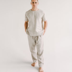 Può includere: Una persona indossa una camicia di lino beige chiaro e pantaloni abbinati. La camicia ha maniche corte e un collo rotondo. I pantaloni hanno una vita con coulisse e una vestibilità rilassata.