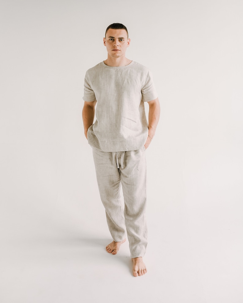 May include: A light beige linen two-piece set. The set includes a short-sleeved, crew neck shirt and drawstring trousers. The fabric has a textured appearance. The model is standing with hands in pockets, showcasing the casual wear.