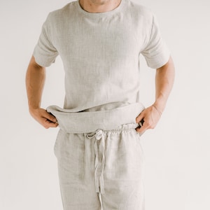 May include: A light grey linen two-piece set. The set includes a short-sleeved crew neck shirt and drawstring trousers. The fabric has a textured appearance. The model is wearing the set. The background is plain white.